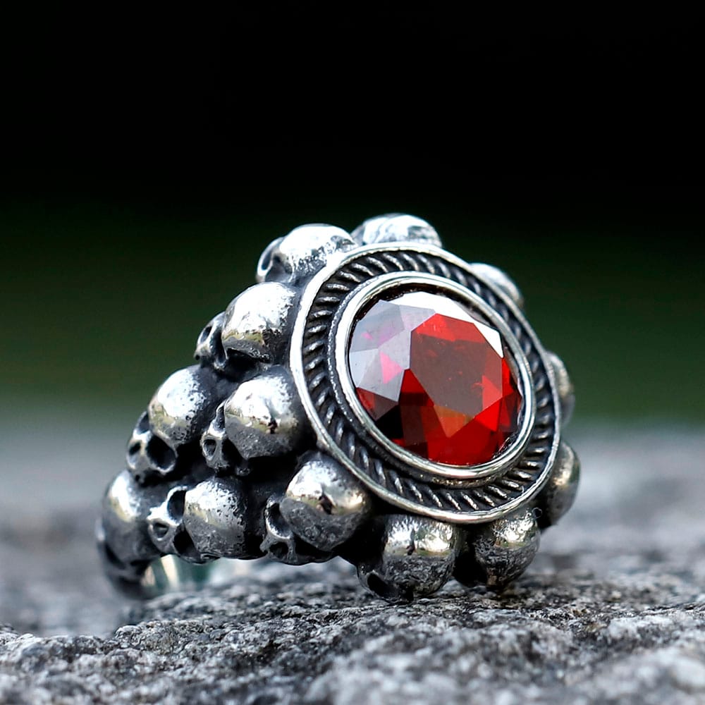 Men's 316L stainless steel skull ring with a large central red zircon stone on a grey rock surface.