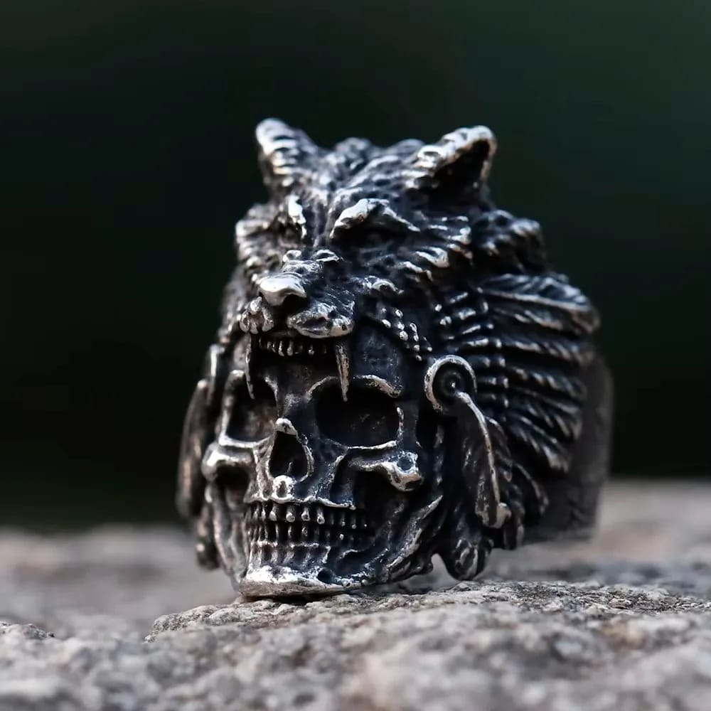 Darkened stainless steel ring featuring a detailed human skull with a wolf head and feathered headdress design, resting on a stone surface.
