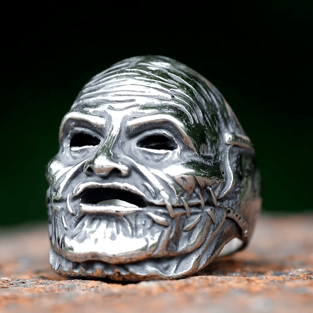 Shiny silver-toned stainless steel ring featuring a detailed gothic mask design with hollow eyes, an open mouth, a wrinkled forehead, and stitched facial details, all against a dark, blurred background.
