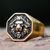 Detailed 3D lion head stainless steel ring, gold tone with black accents, on a rustic dark surface.