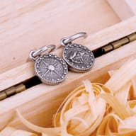 Pair of men's 316L stainless steel dangle earrings with Viking compass and eagle designs on light wood background.