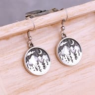 Pair of men's stainless steel drop earrings with mountain forest and moon design, resting on a light wooden surface.