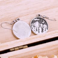 Extreme close-up showcasing the intricate textured mountain and forest design on one round stainless steel earring pendant.