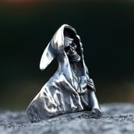 Side view of a gothic antique-finish stainless steel ring featuring a hooded grim reaper skull holding a sickle, on a dark textured surface.