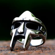 Close-up of the polished stainless steel MF Doom mask ring on a textured dark background.