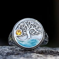 A vintage stainless steel ring with an oxidized Tree of Life design, a yellow zircon, and blue epoxy detail on a textured rock surface.