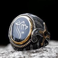 Close-up of a dark, oxidized stainless steel signet ring with a black oval featuring a white Turkish symbol, bordered by gold-colored beads, and a gold-accented sword motif on the side, resting on a stone surface.