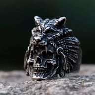 Darkened stainless steel ring featuring a detailed human skull with a wolf head and feathered headdress design, resting on a stone surface.
