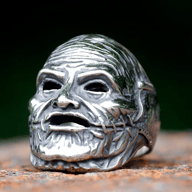 Shiny silver-toned stainless steel ring featuring a detailed gothic mask design with hollow eyes, an open mouth, a wrinkled forehead, and stitched facial details, all against a dark, blurred background.
