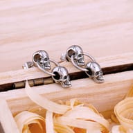 Pair of stainless steel double skull drop earrings resting on a wooden box