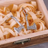 Close-up of the rustic wooden box, partially showing the base of two stainless steel axe drop earrings, on a textured dark surface.