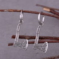 Close-up of two stainless steel Valknut rune axe drop earrings, showing intricate details, with a blurred rustic wooden box in the background.