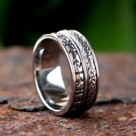 Angled close-up of the stainless steel ring, showcasing both the Celtic knot and rune patterns, resting on a textured rusty surface.