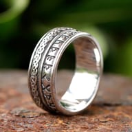 Close-up of a 316L stainless steel ring featuring Elder Futhark runes and Celtic knot patterns, set on a rustic surface with green foliage in the background.