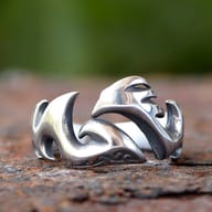Partial close-up of the left side of the flame-inspired stainless steel ring, showing its polished curves against a blurred green background.