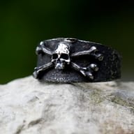 Frontal close-up of the men's skull and crossbones ring, showcasing its wide band and aged finish on wood.