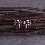 Pair of gothic stainless steel winged skull stud earrings with faceted red gems resting on dark wooden twigs