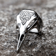 Polished stainless steel ring shaped like a raven's head featuring a prominent Celtic knot on top and smaller knots on the band, resting on a textured gray surface.