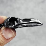 Side profile of the 316L stainless steel raven skull pendant, highlighting the beak and eye socket, held by a finger.