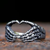 Close-up of a gothic 316L stainless steel ring featuring two skeletal hands forming a heart shape, set on a dark, textured surface.