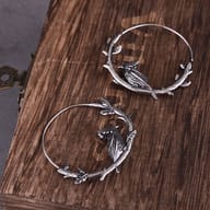 Pair of stainless steel raven on branch hoop earrings displayed on a dark wooden surface