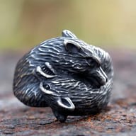A detailed close-up of an antiqued silver-toned metal ring, featuring two intertwined fox heads with textured fur and open mouths, resting on a rusted surface.